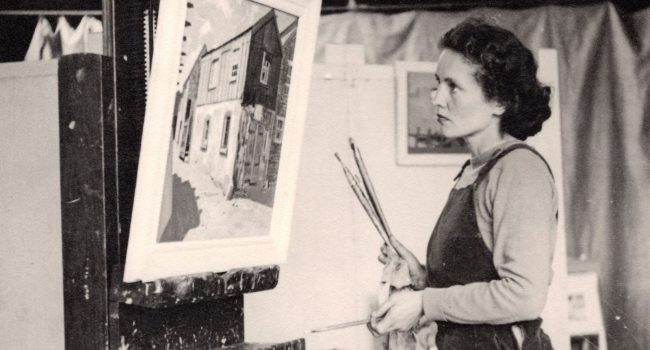 Black and white photograph of Barns-Graham dressed in a dungarees stood in from of a painting of a shed on an easel. She hold several paintbrushes in her right hand