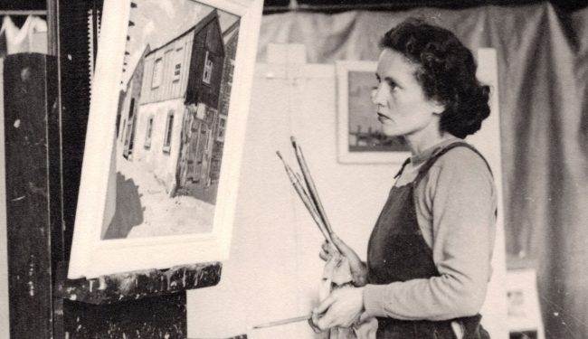 Black and white photograph of Barns-Graham dressed in a dungarees stood in from of a painting of a shed on an easel. She hold several paintbrushes in her right hand