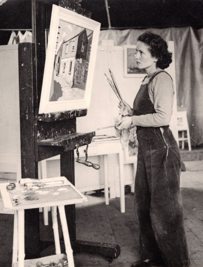 Black and white photograph of Barns-Graham dressed in a dungarees stood in from of a painting of a shed on an easel. She hold several paintbrushes in her right hand