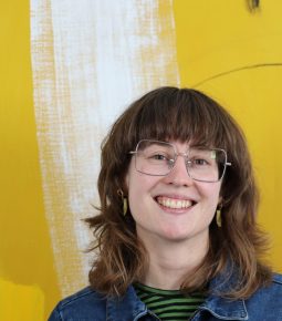 A colour photograph of Tilly Heydon from shoulders up. A yellow painting is behind her