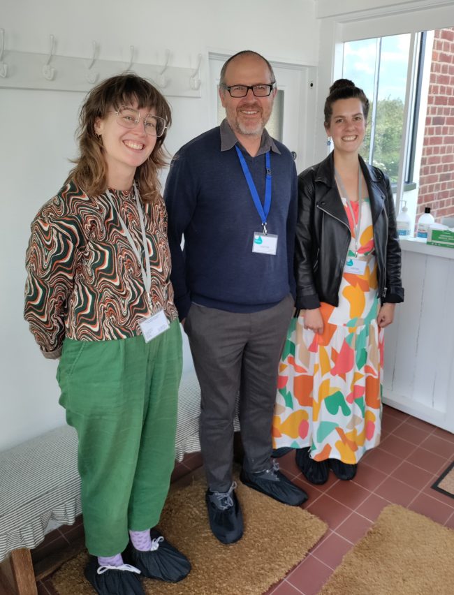 Three people smiling standing in an entrance porch. The porch has white walls, with a coat rack on the wall and the floor is tiled and covered with doormats