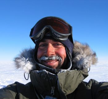Colour photograph of Pete Nienow from shoulders up. He wears a dark green coat with fur edged hood and ski goggles on his head. Snow and blue sky are in the background