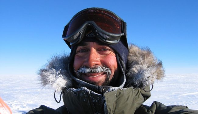 Colour photograph of Pete Nienow from shoulders up. He wears a dark green coat with fur edged hood and ski goggles on his head. Snow and blue sky are in the background