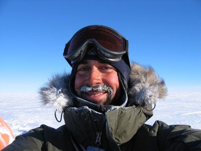 Colour photograph of Pete Nienow from shoulders up. He wears a dark green coat with fur edged hood and ski goggles on his head. Snow and blue sky are in the background