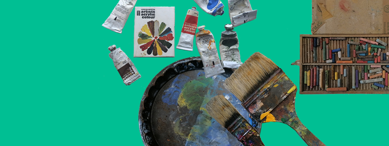 A selection of art materials on an emerald background including tubes of artists acrylic colour and their box, a cake pan with blue and green paint mixed, two wide paintbrushes and a box of pastels.