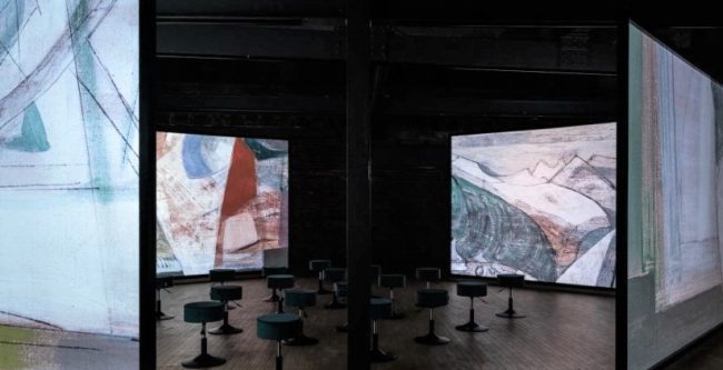 A photo of four screens with close up of abstract paintings projected onto them surrounding stools.