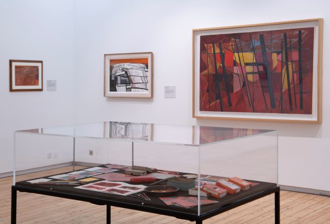 An installation photo of some red abstract artworks with a display case in the foreground with red art materials inside