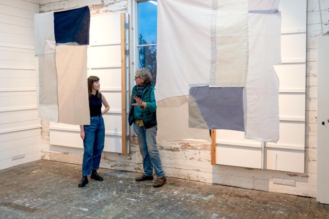 A colour photo of a man and woman talking to each other standing with two fabric hanging partially obscuring them