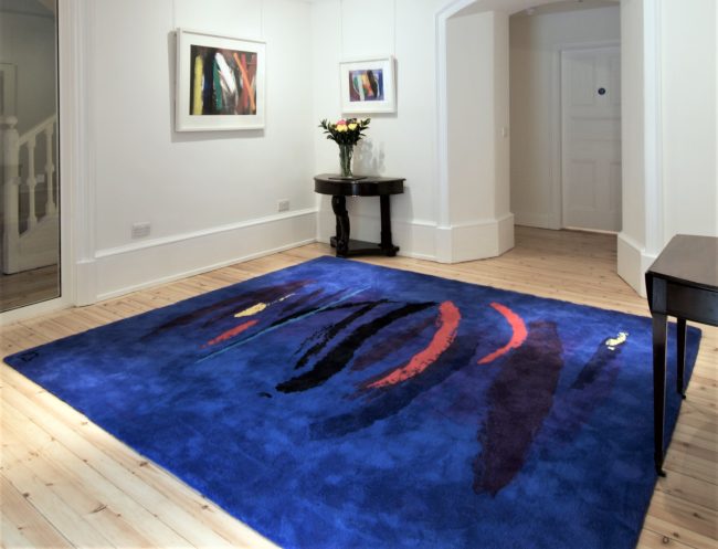 colour photo of blue rug with vertical dashes in black, red and yellow on wooden floor