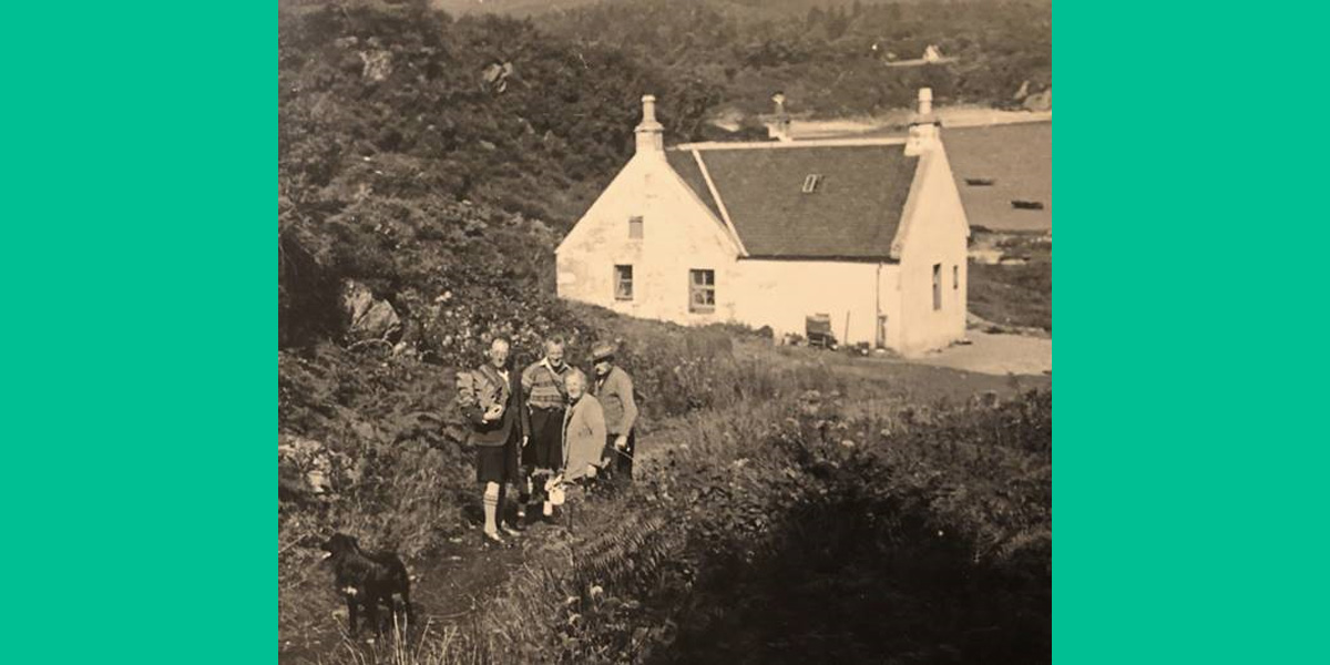 a black and white photograph of a group of 4 people and a black dog standing on a path leading to a white cottage
