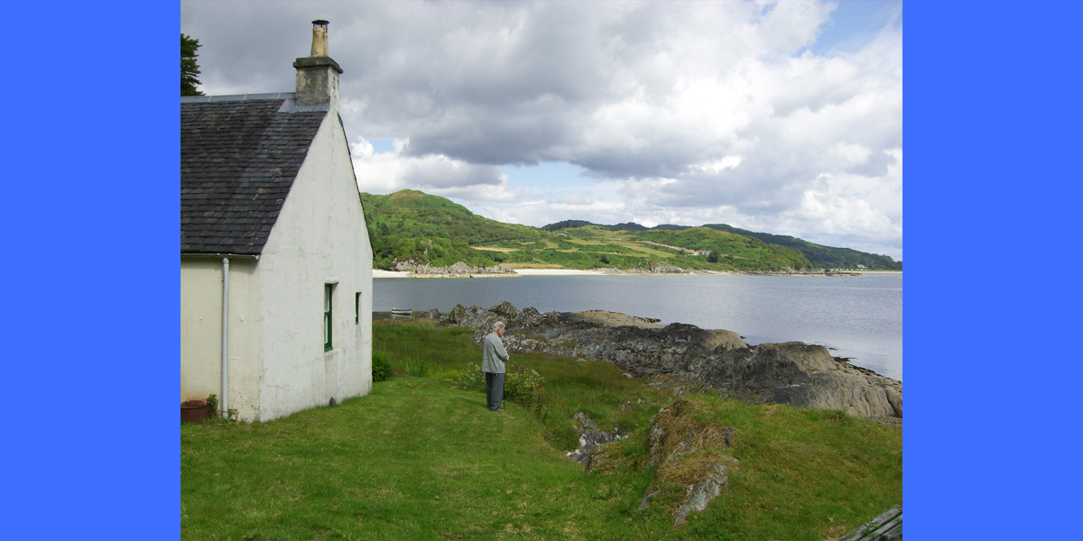 a man standing between a white cottage and the shore of a body of water