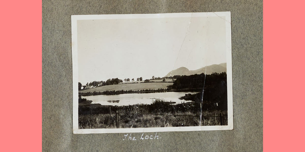 black and white photo of a loch with grass in foreground and hills in far distance. 'The Loch' is written beneath the image