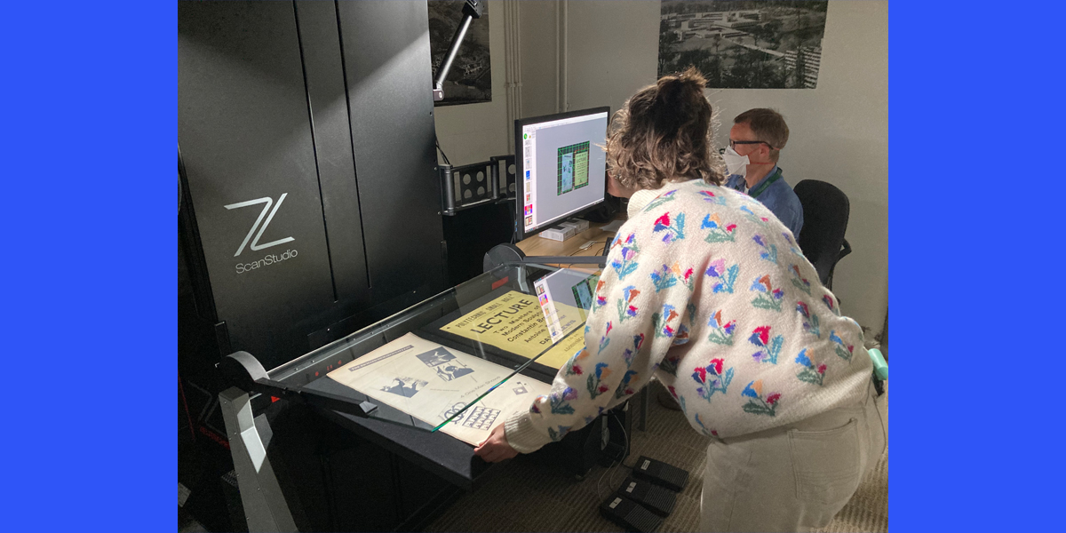 A person standing with a copy stand scanner and a person seated at a computer in the behind