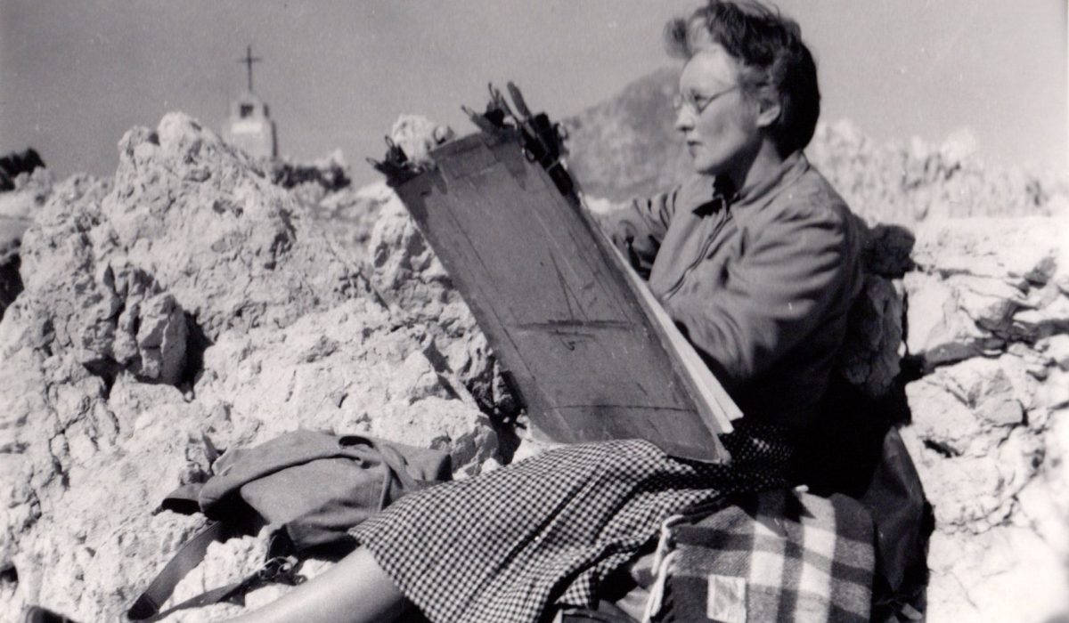 A black and white photo of Barns-Graham seated drawing on a drawing board in a rocky landscape