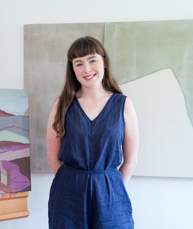 A colour photo of a woman in a blue jumpsuit standing in front of a painting and next to another painting on an easel