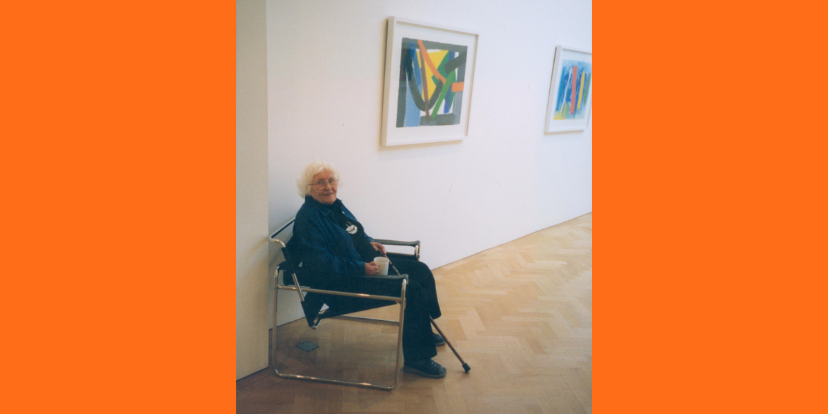 A colour photo of Barns-Graham seated in a chair next to an abstract painting on the wall